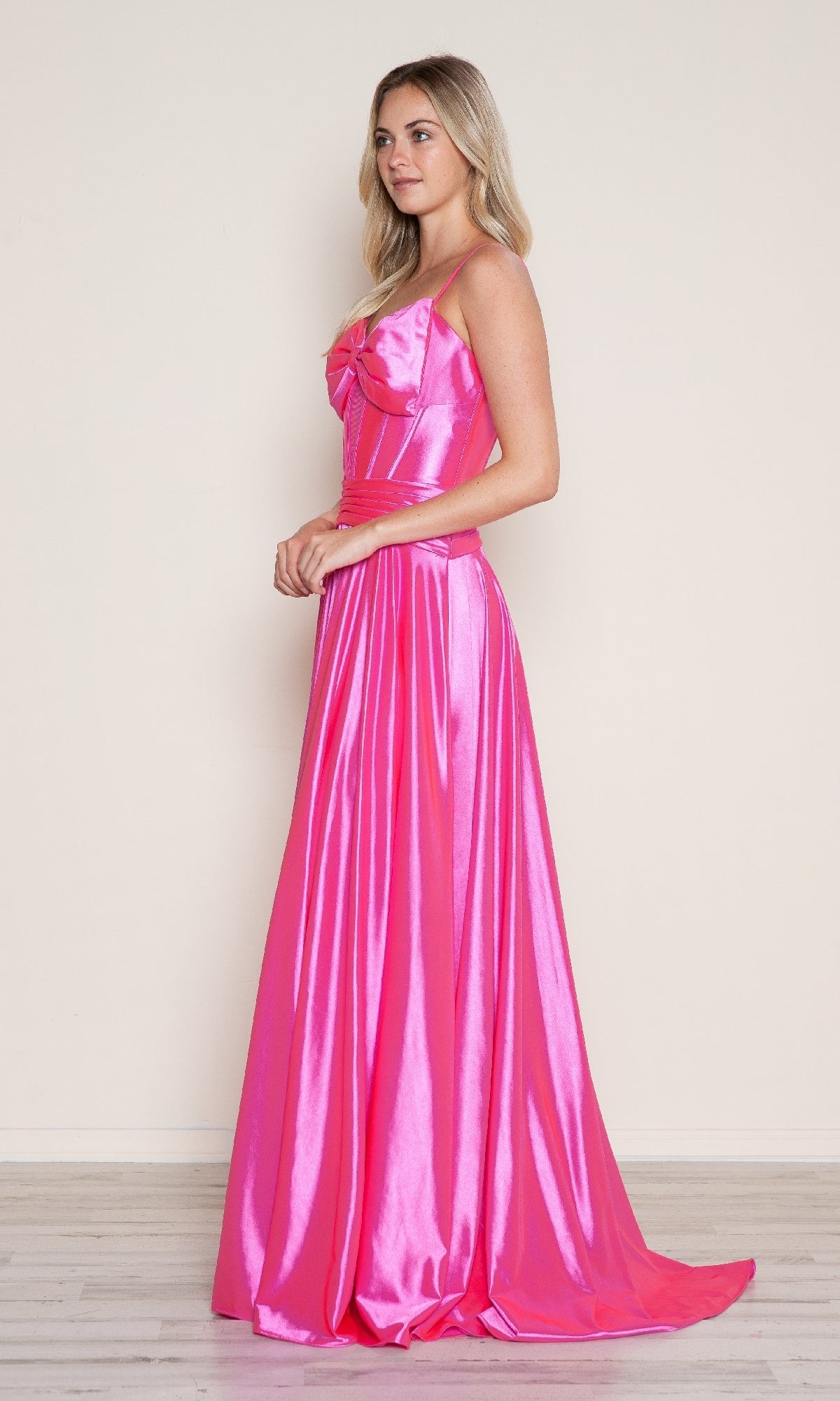 A woman stands in profile against a plain light background wearing the Bow-Front Long Corset Prom Dress: Poly USA 9702, a floor-length hot pink gown featuring a fitted bodice and flared A-line skirt.